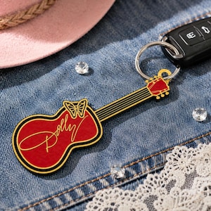 May include: A guitar-shaped keyring featuring a red body with a gold outline and a gold butterfly graphic. The word Dolly is written in gold cursive script across the body. The keyring includes a metal ring attached to a black car key fob.