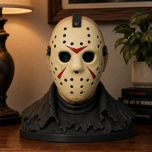 May include: A sculpted bust featuring a cream-colored hockey mask with red chevron markings and multiple ventilation holes, worn over a dark, distressed, textured collar. The mask is secured with a black strap and metal rivets at the forehead.