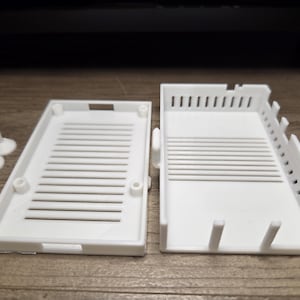 May include: White plastic electronic component case, open to reveal interior. The case has a rectangular shape with a slotted design and several small white plastic feet.