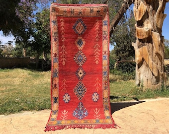 May include: A red, handwoven rug with a geometric pattern in shades of blue, yellow, and white. The rug has fringe on the bottom and is hanging on a wooden post.