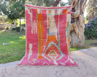 May include: A pink, orange, and green Moroccan rug with a geometric pattern. The rug has a fringe border and is hanging on a wooden post.
