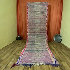 May include: A long, narrow, handwoven rug with a pink, white, and blue color scheme. The rug has a fringe on both ends and a geometric pattern along the edges.