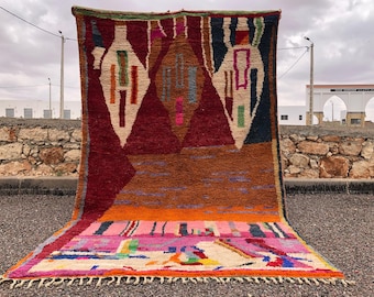May include: A colorful Moroccan rug with geometric patterns in shades of red, orange, pink, blue, and green. The rug has a fringe border and is displayed against a stone wall.