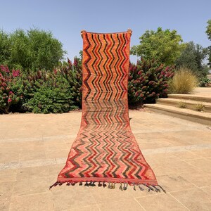 May include: A long, rectangular rug with a zigzag pattern in shades of orange, red, and black. The rug has a fringed edge and is displayed outdoors. The rug's design is a repeating chevron pattern.