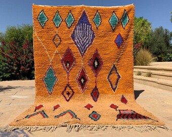 May include: An orange Berber rug with a geometric diamond pattern in teal, red, purple, and black. The rug has a fringed edge and is displayed outdoors. The background includes greenery and a sunny sky.