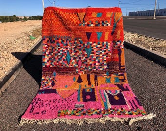 Puede incluir: Una alfombra marroquí colorida con patrones geométricos en tonos de rosa, naranja, azul y verde. La alfombra tiene un borde de flecos y está hecha de lana.