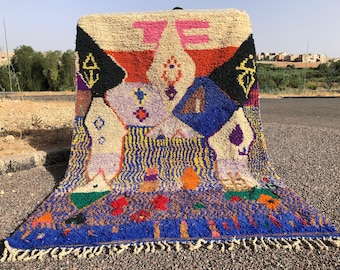 May include: A colorful Moroccan rug with geometric patterns in shades of blue, red, orange, yellow, green, and white. The rug has a fringe border and is made of wool.
