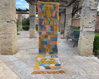 May include: A colorful geometric patterned rug with a fringe border. The rug is made of wool and has a variety of colors, including pink, blue, yellow, and green. The rug is laid out on a stone patio.