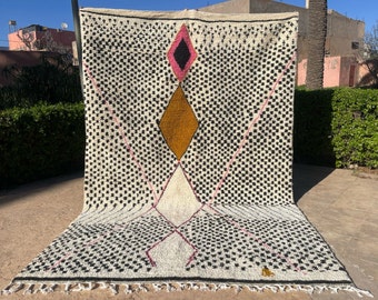 Puede incluir: Una alfombra marroquí blanca, negra, rosa y amarilla mostaza con un patrón geométrico de diamantes. La alfombra tiene un borde de flecos.