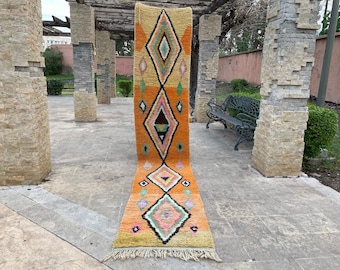 May include: A long, narrow rug with a geometric pattern in shades of orange, yellow, pink, and black. The rug has a fringe on both ends.