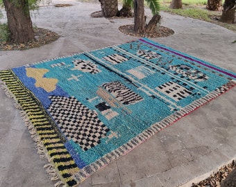 May include: A colourful Moroccan rug with a blue background and geometric patterns in black, white, yellow, and red. The rug has a fringed border.