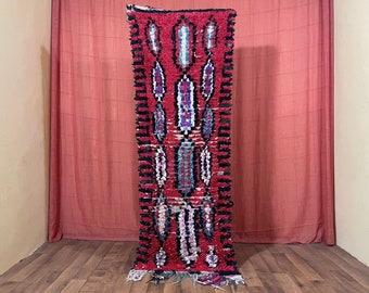Puede incluir: Una alfombra marroquí roja, negra y azul con un patrón geométrico. La alfombra está tejida a mano y tiene flecos en los extremos.