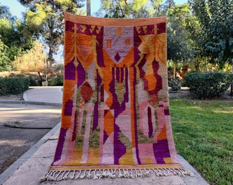 May include: A colorful Moroccan rug with geometric patterns in shades of orange, pink, purple, and green. The rug has a fringe border and is hanging on a wall.