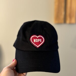 Könnte beinhalten: Schwarze Baseballkappe mit einem roten Herz-Patch, auf dem "NOPE" in weißen Buchstaben steht.