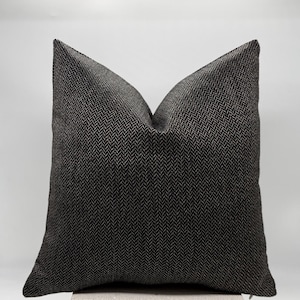 May include: A square decorative pillow with a dark gray and black herringbone pattern. The pillow is resting on a light gray surface and has a pointed top.