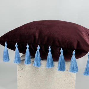 Puede incluir: Un cojín cuadrado hecho de tela de terciopelo color burdeos intenso. Los bordes del cojín están decorados con una hilera de borlas de color azul claro que cuelgan de las costuras, creando un contraste marcado con la textura suave del terciopelo.