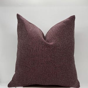 May include: A square cushion with a herringbone pattern in shades of burgundy and dark red. The cushion is placed on a light-coloured surface against a white background. The cushion has a zip closure.