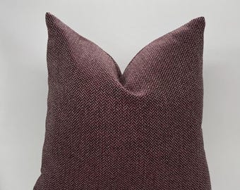 May include: A square decorative pillow with a herringbone pattern in shades of burgundy and dark red. The pillow is placed on a light-colored surface against a white background. The pillow has a zipper closure.