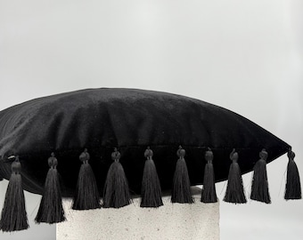 May include: A black velvet pillow with a row of black tassels along the bottom edge. The pillow is resting on a light-colored, textured surface. The pillow is a decorative home accent.