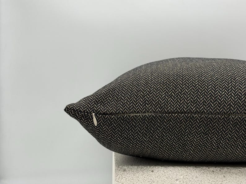 May include: Close-up of a decorative pillow with a black and white herringbone pattern. The pillow has a zipper closure and is resting on a light-colored surface. The pillow is a neutral color and would complement many decor styles.