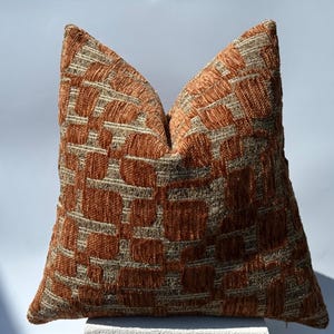 May include: A square throw cushion featuring a textured, raised geometric pattern in burnt orange and metallic gold. The design consists of irregular, block-like shapes woven into a dense, tactile surface, creating a distinct contrast between the colours.