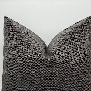 May include: A decorative pillow with a dark gray and beige herringbone pattern. The pillow has a soft, textured appearance and a classic design. The pillow is a square shape and is suitable for a bed or sofa.
