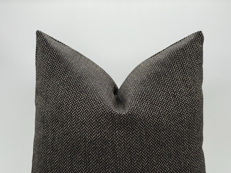 May include: A decorative pillow with a dark gray and beige herringbone pattern. The pillow has a soft, textured appearance and a classic design. The pillow is a square shape and is suitable for a bed or sofa.