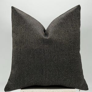 May include: A square decorative pillow with a dark gray and beige herringbone pattern. The pillow has a soft, textured appearance and is designed for home decor. The pillow is approximately 18 inches square.
