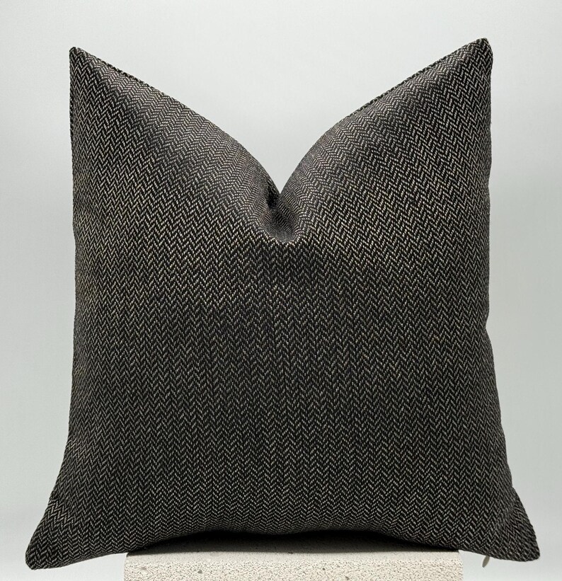 May include: A square decorative pillow with a dark gray and beige herringbone pattern. The pillow has a soft, textured appearance and is designed for home decor. The pillow is approximately 18 inches square.