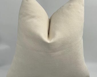 May include: A cream-colored throw pillow with a subtle textured fabric. The pillow is sitting on a white block.