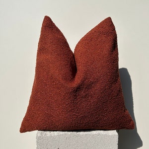 May include: A textured, rust-colored decorative pillow. The pillow is square-shaped and has a soft, plush appearance. It is placed on a white, rectangular block against a white background. The pillow's color is a warm, earthy tone.