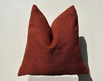 Op de afbeelding: Een vierkant, roestkleurig decoratief kussen met een getextureerd oppervlak. Het kussen heeft een zachte, pluche uitstraling. Het is geplaatst op een witte, rechthoekige blok tegen een witte achtergrond. De kleur is een warme, aardse tint.