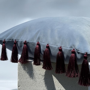 May include: A square throw pillow in a solid dusty blue color featuring a smooth, soft-touch fabric finish. The bottom edge of the pillow is decorated with a row of nine deep burgundy red tassels that hang vertically from the seam.