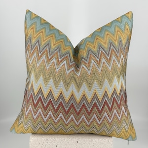 May include: A square throw pillow featuring a repeating chevron pattern in shades of light blue, mustard yellow, metallic gold, and deep red. The fabric has a textured, woven appearance with a shimmering finish throughout the zigzag design.