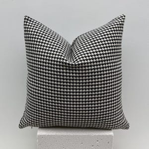 Könnte beinhalten: Ein quadratisches Zierkissen mit einem schwarz-weißen Hahnentrittmuster. Das Kissen besteht aus einem weichen Stoff und ist mit einem flauschigen Material gefüllt. Das Kissen liegt auf einer weißen Oberfläche.