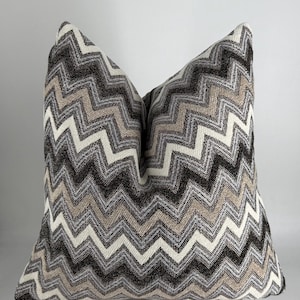 May include: A cushion featuring a zig-zag pattern in black, white, and beige tones. The square cushion has a textured fabric appearance. The design is a repeating chevron pattern, suitable for home decor.