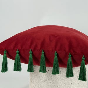 May include: A square throw pillow made of deep red velvet fabric. The edges of the pillow are decorated with a series of hanging dark green tassels that are evenly spaced along the bottom seam, creating a distinct contrast in color and texture.