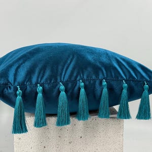 Puede incluir: Cojín de terciopelo color teal con borlas teal a lo largo del borde inferior. El cojín es de color teal liso y parece suave y afelpado. Las borlas están espaciadas uniformemente y añaden un elemento decorativo.