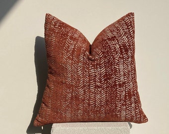 Peut inclure: Un coussin de décoration de couleur rouille avec un motif en chevron blanc. Le coussin est posé sur un piédestal blanc.
