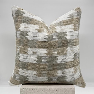 May include: A square throw pillow with a geometric pattern in shades of beige, white, and gray. The pillow is sitting on a light gray block.