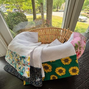 May include: A wicker basket with a white interior, lined with patterned fabric. The fabric features sunflowers, floral patterns, and decorative motifs. The basket sits on a dark surface, with a bright outdoor scene visible in the background. The basket is made of woven wicker.