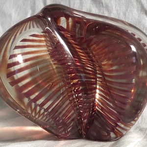 May include: A clear glass sculpture with a rounded, organic shape. The interior features intricate, reddish-brown linear patterns that create a striped effect. The glass is smooth and reflects light, highlighting the internal design.