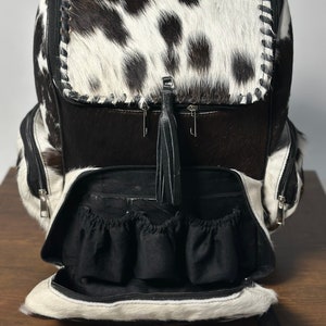 May include: A black and white cowhide backpack with a black leather tassel. The backpack has multiple pockets and is open to reveal a black fabric lining with several smaller pockets.
