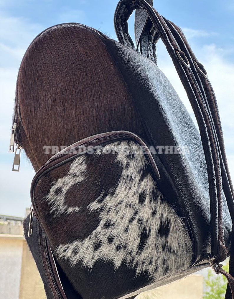 Pu&ograve; includere: Uno zaino in pelle marrone con una toppa in pelle di mucca bianca e marrone sul davanti. Lo zaino ha una tasca con cerniera sul davanti e due spalline regolabili.