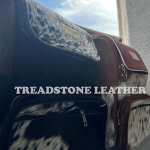 Pu&ograve; includere: Due zaini in pelle marrone con dettagli in pelle di mucca bianca e nera. Gli zaini hanno una chiusura a zip e il testo "TREADSTONE LEATHER" &egrave; stampato sulla parte anteriore di uno degli zaini.