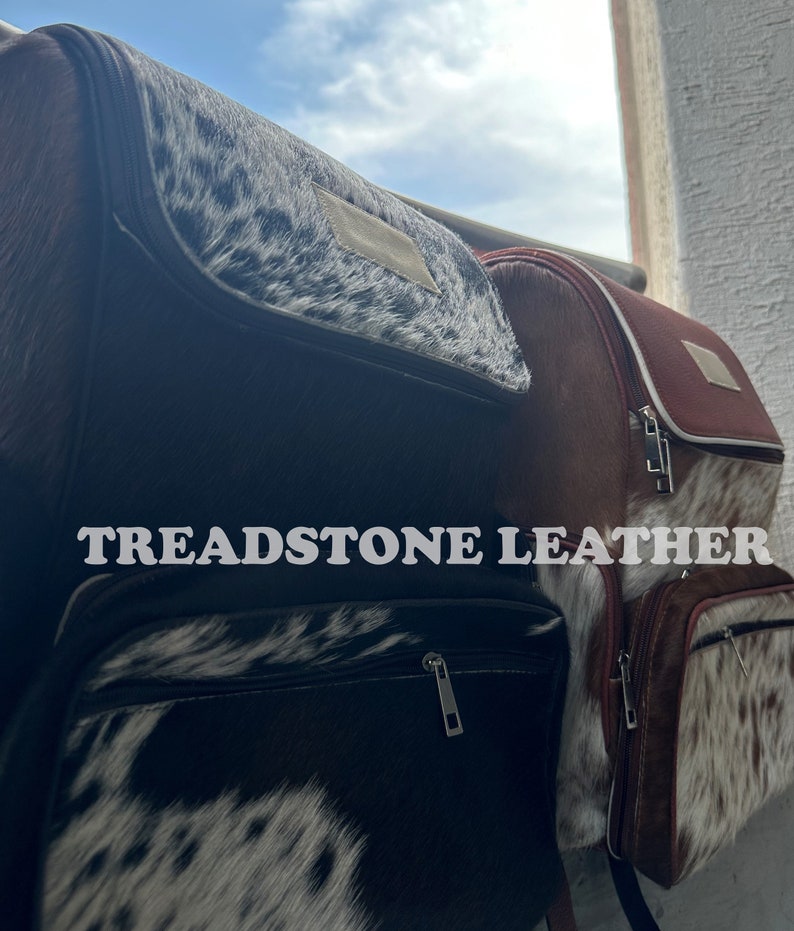 Pu&ograve; includere: Due zaini in pelle marrone con dettagli in pelle di mucca bianca e nera. Gli zaini hanno una chiusura a zip e il testo "TREADSTONE LEATHER" &egrave; stampato sulla parte anteriore di uno degli zaini.
