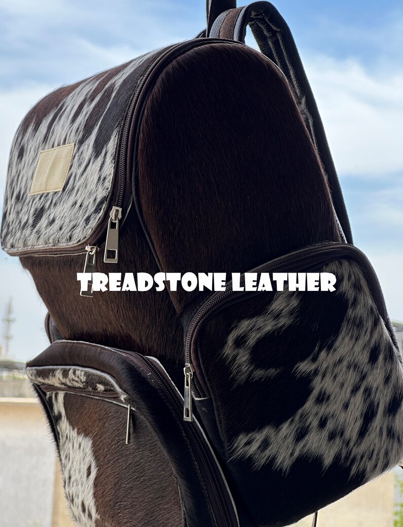 Pu&ograve; includere: Uno zaino in pelle marrone con un motivo in pelle di mucca bianca e marrone. Lo zaino ha un grande scomparto principale e una tasca anteriore pi&ugrave; piccola. Il testo "TREADSTONE LEATHER" &egrave; stampato sulla parte anteriore dello zaino.