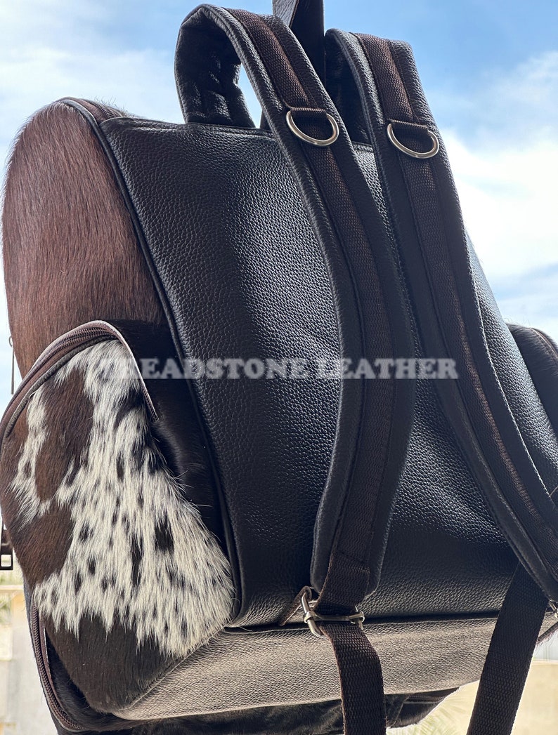 Pu&ograve; includere: Zaino in pelle marrone con una toppa in pelle di mucca sul davanti. Lo zaino ha due spalline regolabili e una tasca con cerniera sul davanti. Il testo "Headstone Leather" &egrave; visibile sul retro.