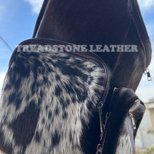 Pu&ograve; includere: Uno zaino in pelle marrone con un motivo in pelle di mucca bianca e nera sul lembo anteriore. Lo zaino ha una chiusura a zip e un logo Treadstone Leather.