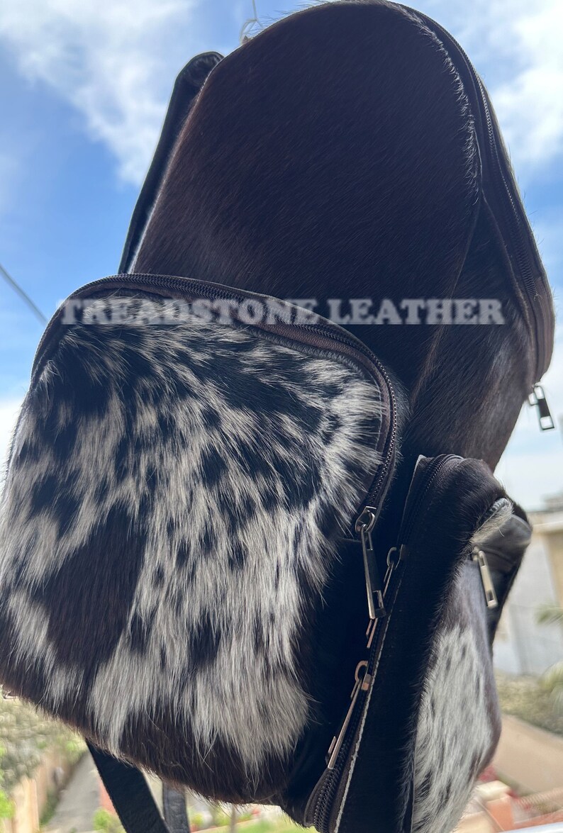 Pu&ograve; includere: Uno zaino in pelle marrone con un motivo in pelle di mucca bianca e nera sul lembo anteriore. Lo zaino ha una chiusura a zip e un logo Treadstone Leather.